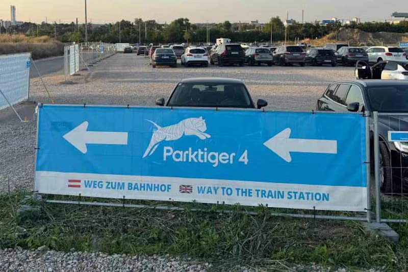 Blaues Banner mit Parktiger 4 Logo und Pfeilen vor kiesigem Parkplatz mit geparkten Autos und Bauzaun.