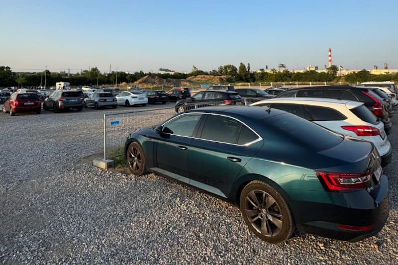 Dunkelgrünes Auto im Vordergrund auf geschottertem Parkplatz, Reihen geparkter Fahrzeuge und Zaun im Hintergrund.