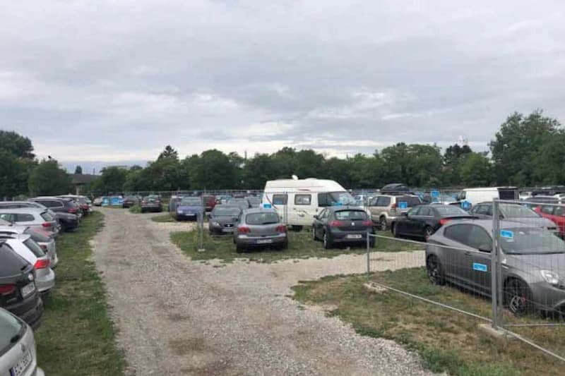 Geschotterter Parkplatz mit Bauzäunen, zahlreichen Autos und einem Wohnmobil vor bewölktem Himmel.