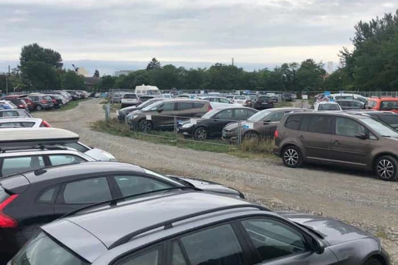 Geschotterter Parkplatz mit dicht geparkten Autos, Drahtzaun und Bäumen unter bewölktem Himmel.
