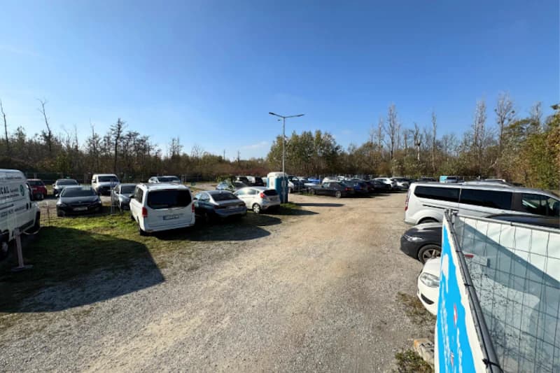 Schotterparkplatz mit zahlreichen geparkten Autos, mobiler Toilette, Beleuchtungsmast und Bauzaun rechts, Baumreihe im Hintergrund.