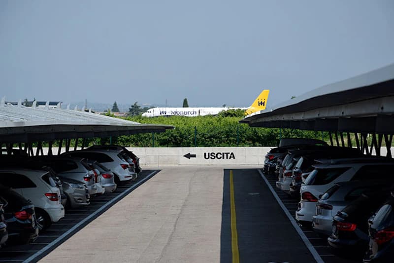 Überdachter Parkplatz mit zwei Reihen geparkter Autos, mittlere Fahrspur und Wand mit Pfeil und Aufschrift USCITA; Flugzeugheck im Hintergrund.