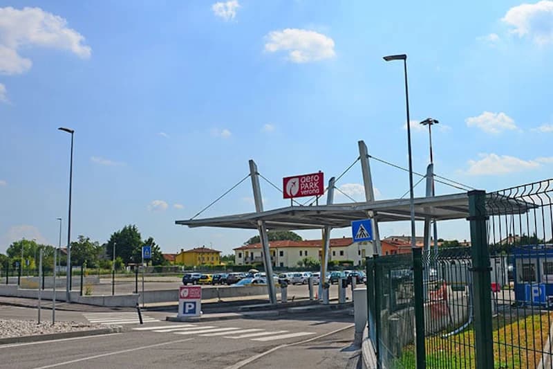 Eingang mit überdachtem Tor, Schild Aeropark Verona, umzäunter Parkplatz und parkende Autos.