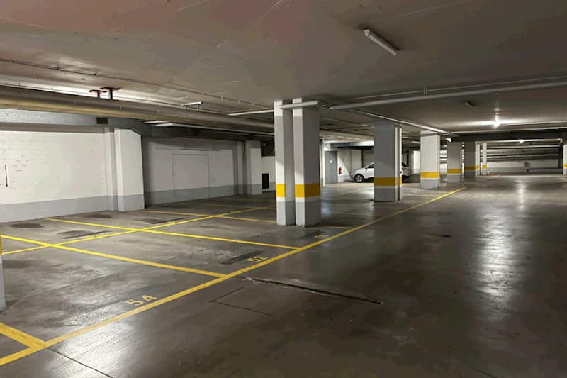 Überdachtes Parkhaus mit markierten Parkfeldern, Stützpfeilern und einem abgestellten Wagen im Hintergrund.