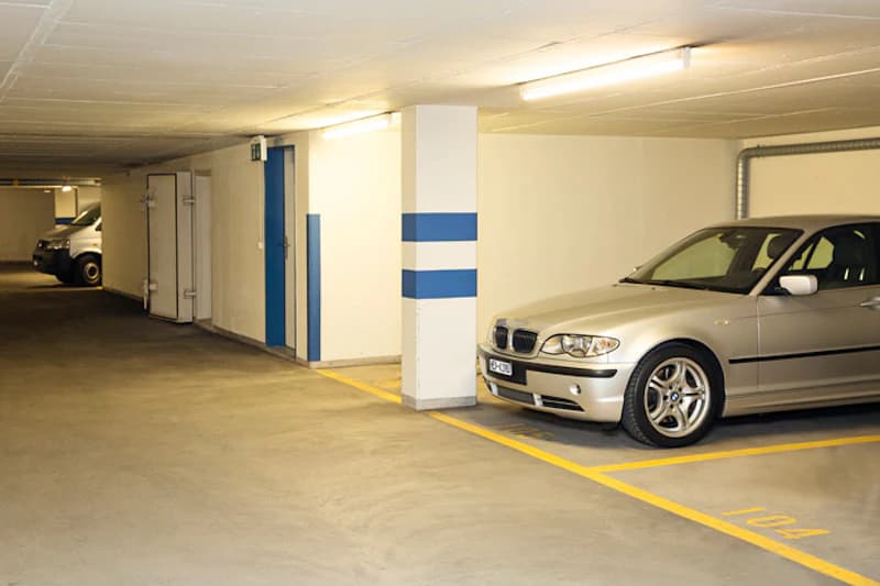 Überdachter Parkdeckgang mit silbernem Auto auf markiertem Stellplatz neben Säule mit blauen Streifen.