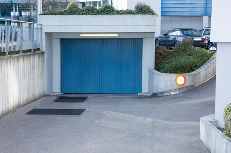 Überdachte Garage mit geschlossenem blauen Tor, beleuchteter Einfahrt und Zufahrtsrampe.