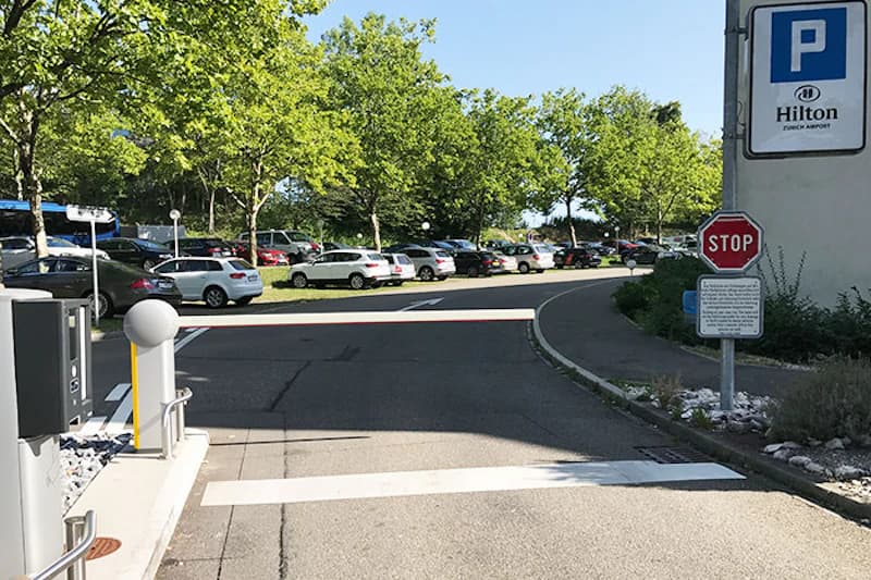 Einfahrtschranke vor offenem Parkplatz mit parkenden Autos, Stoppschild und Hilton-Parkschild.
