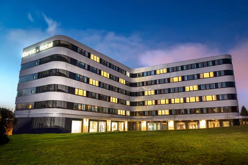 Mehrstöckiges Hotel mit geschwungener Fassade und zahlreichen beleuchteten Fenstern bei Dämmerung, Rasen im Vordergrund.