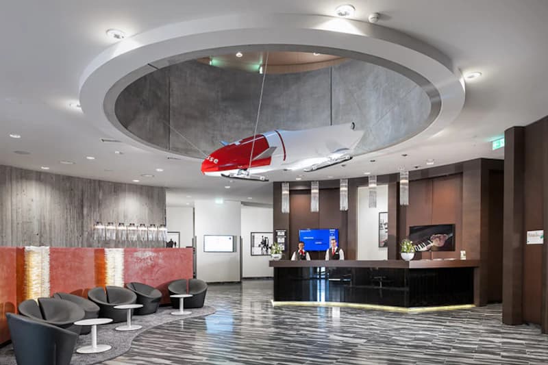 Hotelhalle mit Empfangstheke, Sitzgruppe links und hängendem Modellflugzeug unter rundem Deckenausschnitt.