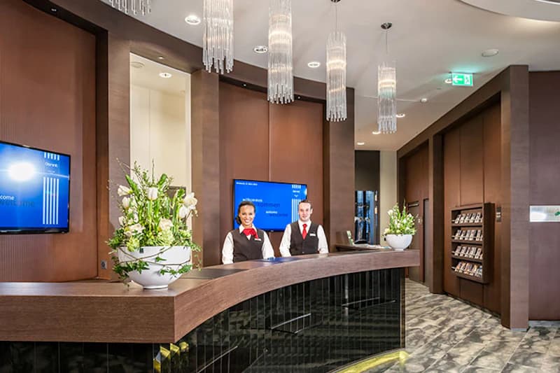 Hotelrezeption mit geschwungenem Holztresen, zwei Empfangsmitarbeitenden in Uniform, Blumenarrangements und Kronleuchtern.
