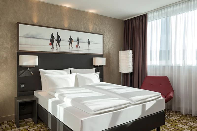 Hotelzimmer mit Doppelbett, zwei Nachttischen, Wandlampen, großem Bild, Stehlampe und rotem Sessel am Fenster.