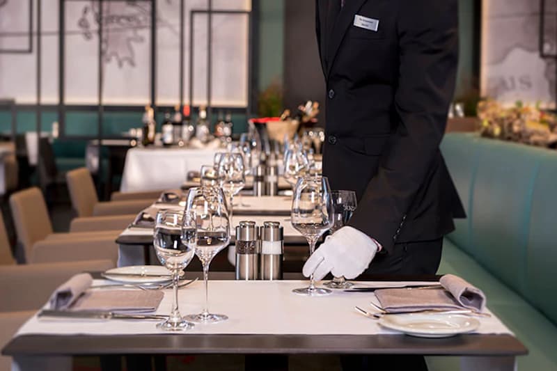 Gepflegte Tischreihe im Hotelrestaurant, Servicekraft mit weißen Handschuhen richtet ein Weinglas.