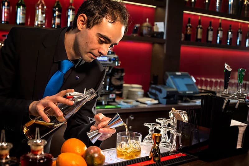 Barkeeper gießt eine Spirituose mit Messbecher in ein Glas an einer Hotelbar, Orangen und Glasware im Vordergrund.