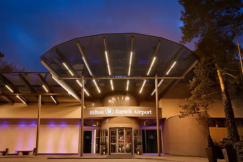 Eingangsbereich des Hilton Zurich Airport bei Dämmerung mit beleuchtetem Vordach und Drehtür.