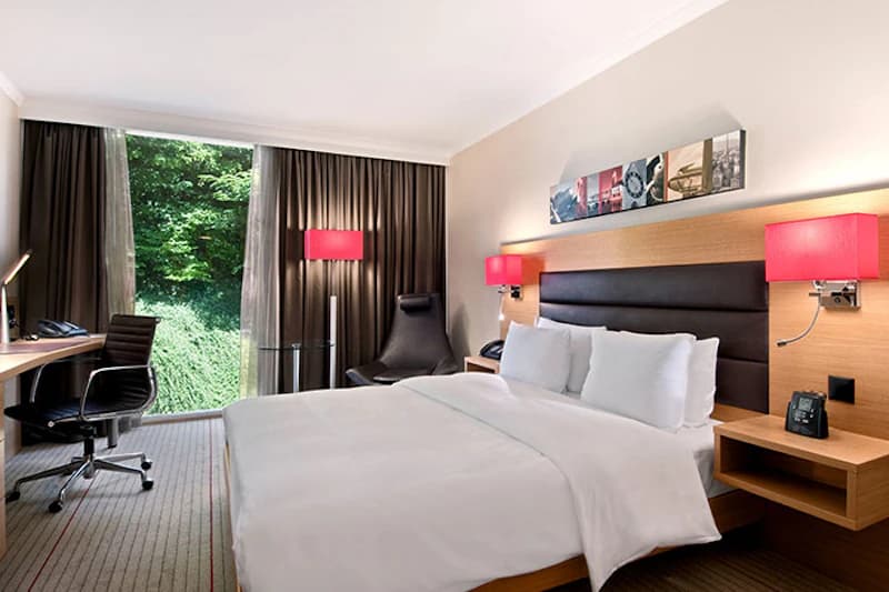 Doppelzimmer mit großem Bett, roten Wandlampen, Schreibtisch mit Stuhl, Sessel und großem Fenster zum Grün.</