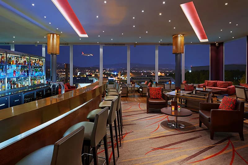 Hotelbar mit Sitzgruppen, Theke und großen Fenstern, Nachthimmel mit anfliegendem Flugzeug und Stadtlichtern.