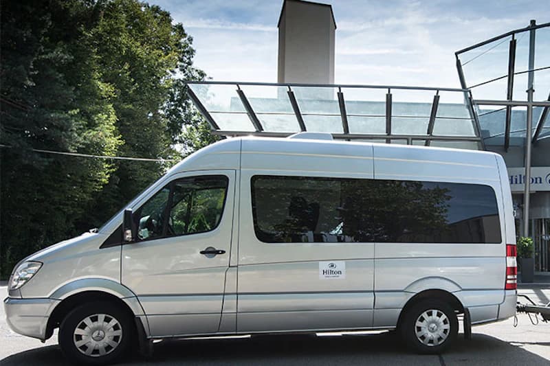 Silberner Kleinbus mit Hilton-Logo steht vor Glasschutzdach am Hoteleingang, Bäume im Hintergrund.
