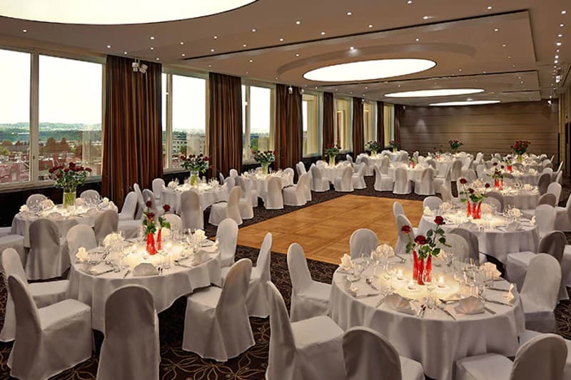 Bankettsaal mit eingedeckten runden Tischen, weißen Stuhlhussen, roten Blumen und zentraler Tanzfläche vor großen Fenstern.