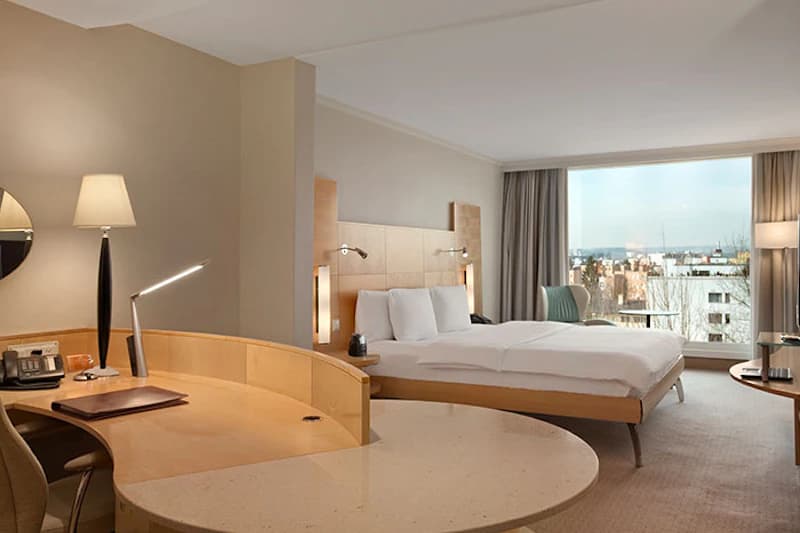 Hotelzimmer mit Doppelbett, Schreibtisch, Lampen und grossem Fenster mit Stadtblick.