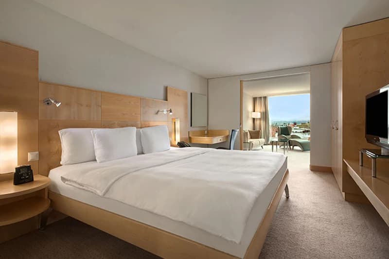 Helles Hotelzimmer mit großem Bett, Holzkopfteil, Nachttischen, Schreibtisch, Fernseher und Balkon mit Sitzbereich.