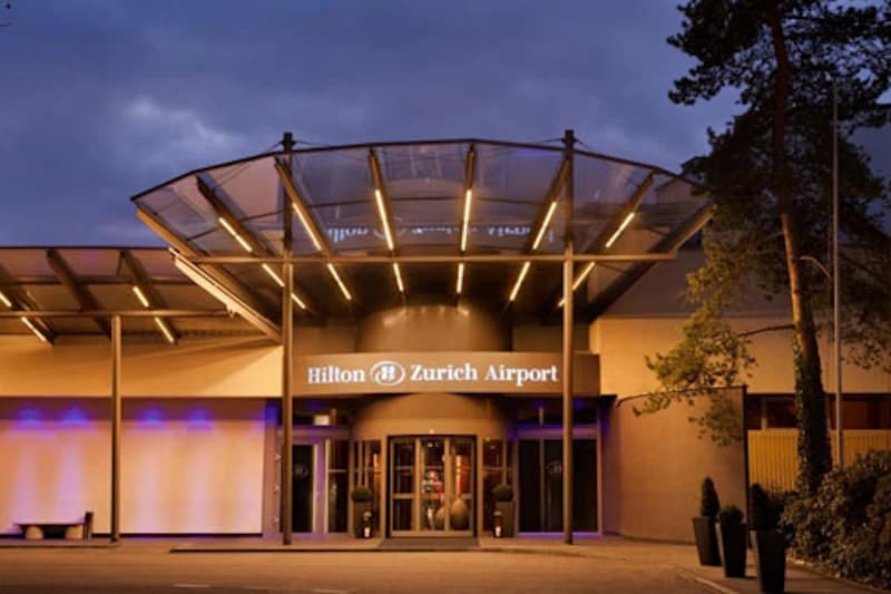 Eingangsportal des Hilton Zurich Airport mit beleuchtetem Vordach und Drehtür bei Dämmerung.
