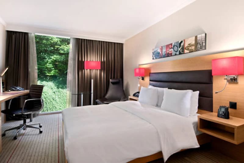 Hotelzimmer mit Doppelbett, gepolstertem Kopfteil, Nachttischen und großem Fenster mit Blick ins Grüne.