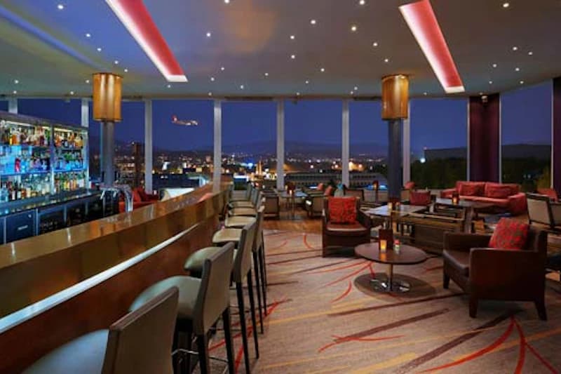 Hotelbar und Lounge mit Bartresen, Sitzgruppen und grossen Fensterfronten zum nächtlichen Flughafenblick.