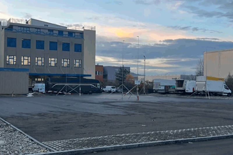 Leerer asphaltierter Parkplatz vor Industriegebäuden mit geparkten Lieferwagen und Anhängern unter bewölktem Himmel.