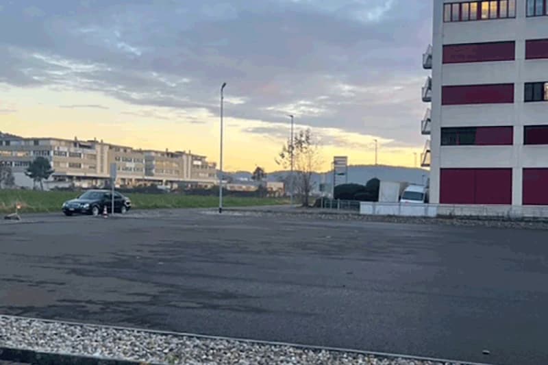 Leerer Asphaltparkplatz, links ein schwarzes Auto, Laternen und Wohngebäude im Hintergrund bei Abenddämmerung.
