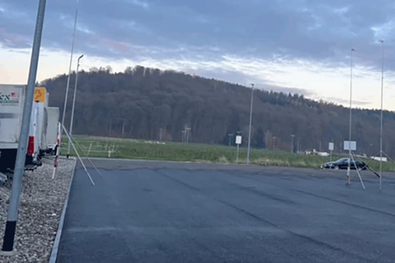 Leerer Parkplatz mit Lieferwagen links, einem Auto rechts, Lichtmasten und bewaldetem Hügel im Hintergrund.
