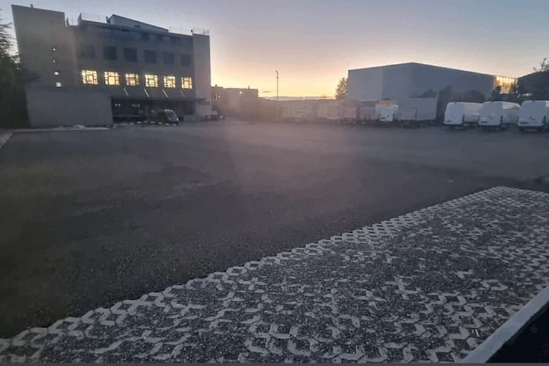 Große gepflasterte Freifläche bei Dämmerung, im Vordergrund strukturierte Rasengitterplatten, links Gebäude mit beleuchteten Fenstern, rechts Lieferwagen.