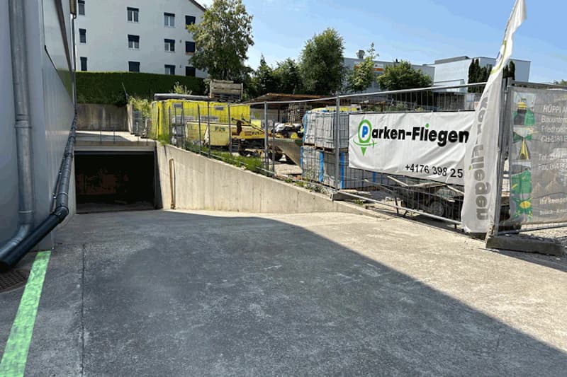 Absteigende Zufahrt zur Tiefgarage mit Betonrampe, Zaun mit Parken-Fliegen-Banner und Baustelle rechts.