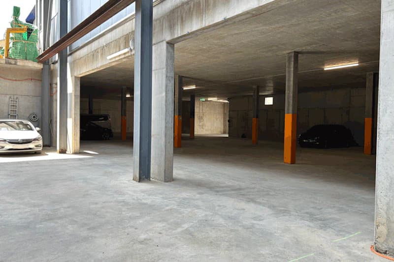 Betonierte Tiefgarage mit Stützsäulen, orange Schutzummantelungen, einigen geparkten Autos und beleuchtetem Innenraum.