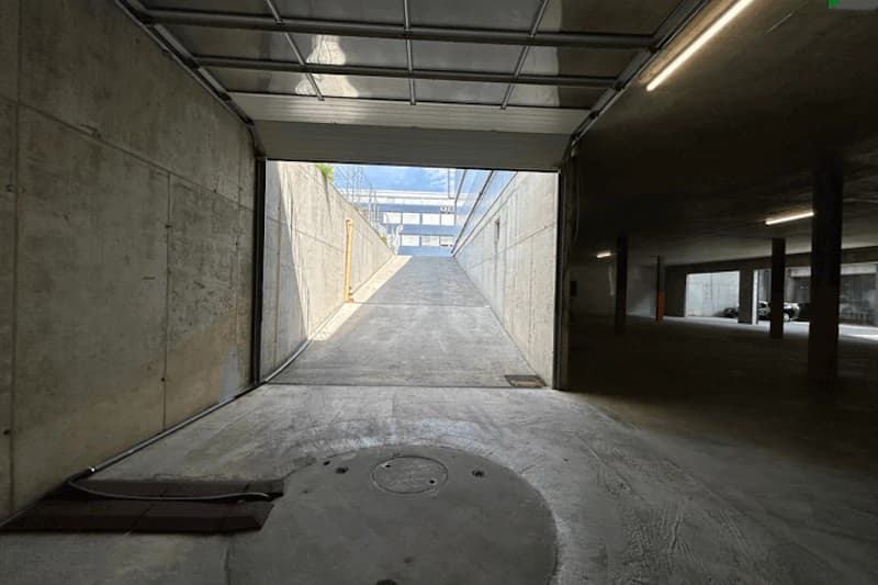 Tiefgaragenausfahrt mit Rampe zu Tageslicht, offene Rolltür, Betonwände und beleuchteter Innenbereich mit Stützen.