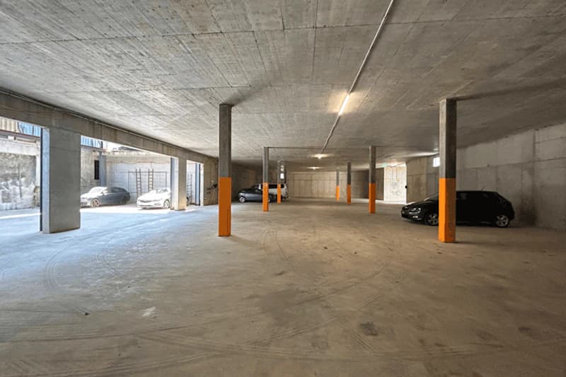 Tiefgarage mit Betonstützen, orange markierten Säulen, Deckenbeleuchtung und wenigen geparkten Autos.