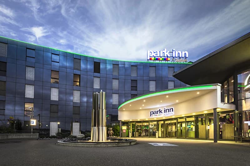 Abendaufnahme des beleuchteten Hoteleingangs Park Inn mit rundem Vorplatz und Metallskulptur.