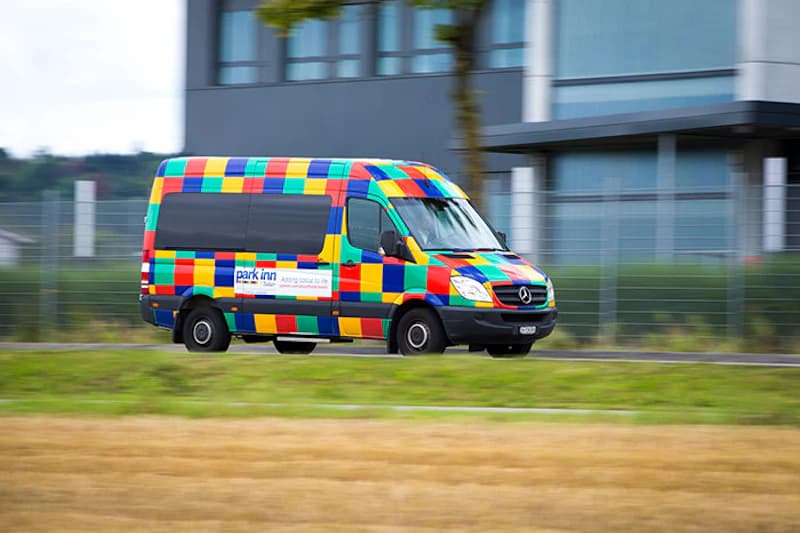 Bunter karierter Shuttlebus mit Park Inn Schriftzug fährt vor Gebäuden am Flughafen Zürich.