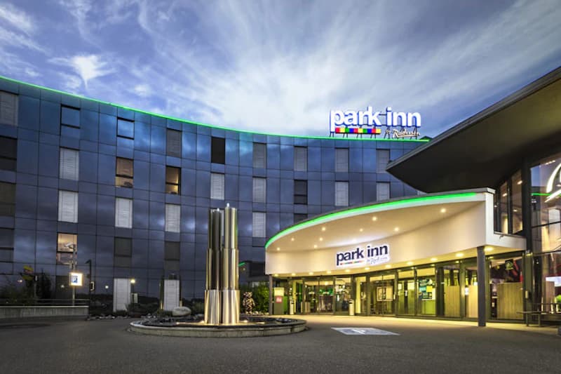 Park Inn Hoteleingang mit beleuchtetem Vordach, rundem Brunnen und mehrstöckiger Fassadenfront bei Dämmerung.